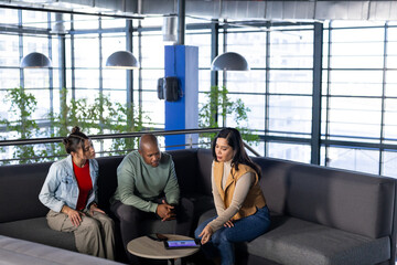 Diverse coworkers sitting on sofa in office lounge discussing tablet on coffee table with plants