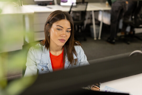 Indian woman wearing denim jacket holding chart and analyzing data at office desk with computer