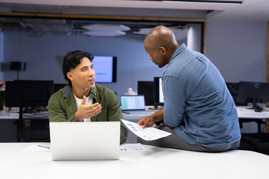 Diverse male coworkers discussing project data in open-plan office with laptop and printed charts