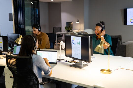 Diverse support staff collaborating at white desk in open-plan office using headsets and monitors