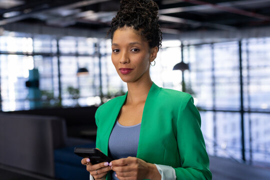 African American woman standing in office lounge holding smartphone wearing green blazer