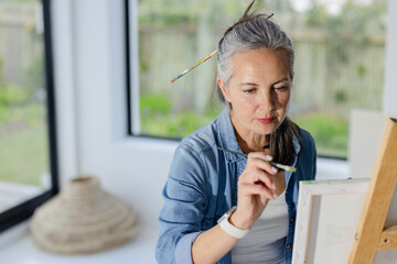 Senior woman painting on canvas at easel in art studio near black-framed windows wearing smartwatch
