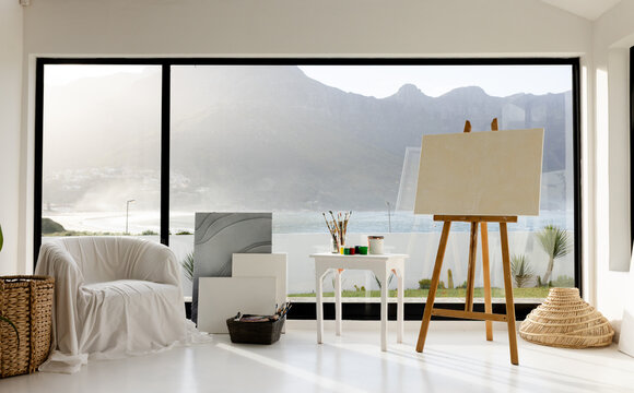 Wooden easel holding blank canvas by table with brushes at windows overlooking lake and mountains
