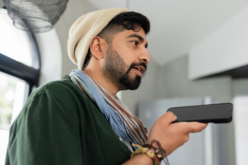 Indian man holding smartphone near mouth while speaking in modern kitchen with refrigerator