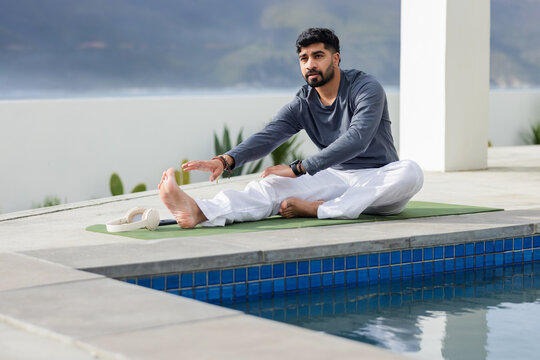 Adult Indian man sitting on yoga mat stretching leg by blue pool with white headphones