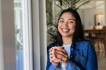 Woman holding ceramic coffee mug and smiling beside large glass window and potted plant in cafe
