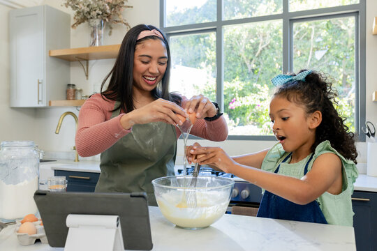Hispanic mother and daughter cracking egg into bowl, whisking batter with tablet at kitchen island - Powered by Adobe