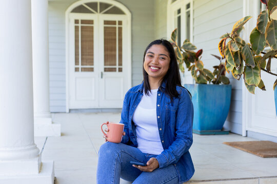 Asian woman sitting on front porch steps holding coral-colored mug and checking smartphone