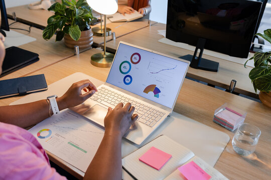White laptop is displaying colorful charts and is resting on wooden desk with notebooks and plants