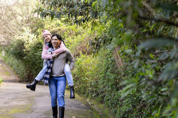 Diverse couple walking wooded path carrying woman on back wearing pink quilted jacket and beanie
