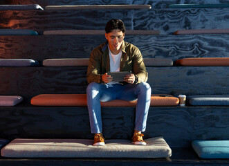 Hispanic male student sitting on bleachers holding tablet with coffee cup beside bench cushions