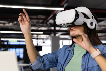 Asian woman wearing denim shirt, white VR headset reaching in office using laptop and smartwatch