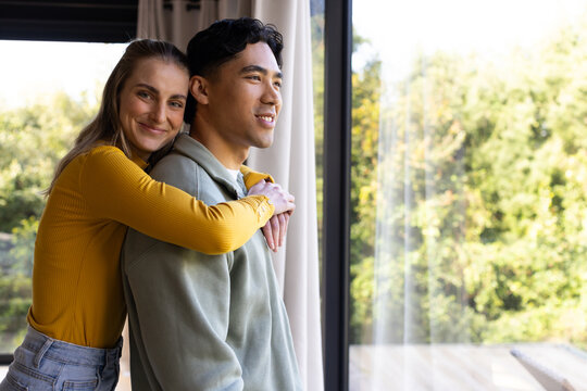 Asian man and woman embracing, gazing through curtains by sliding glass door onto deck, copy space - Powered by Adobe