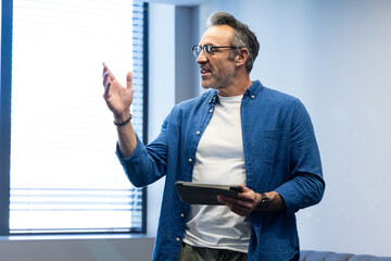 Man holding tablet and gesturing with right hand in office near window blinds wearing denim shirt