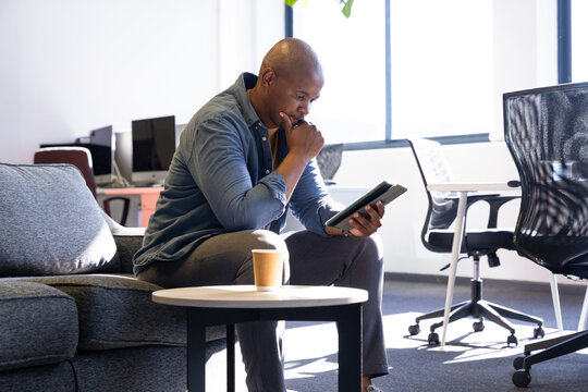 African American man using tablet and sipping cup on gray sofa beside table in office lounge