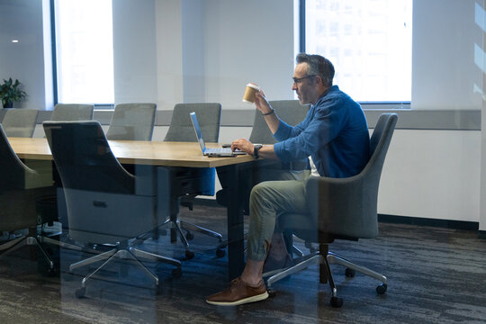 Laptop resting on conference table with empty swivel chairs and potted plant near cityscape windows