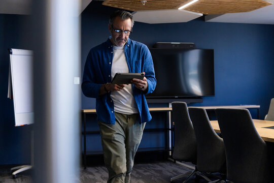 Man standing in conference room in business casual attire holding tablet near flat-screen display