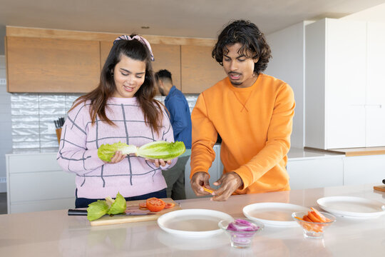 Friends prepping at kitchen island, woman holding romaine in pink check sweater, man slicing pepper