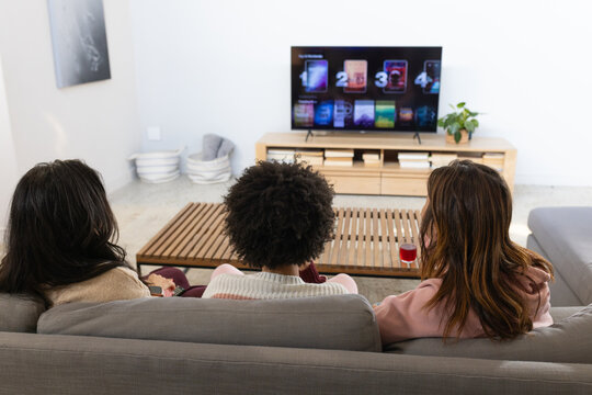 Diverse female friends sitting on gray sofa in living room, holding remote and drink, watching TV