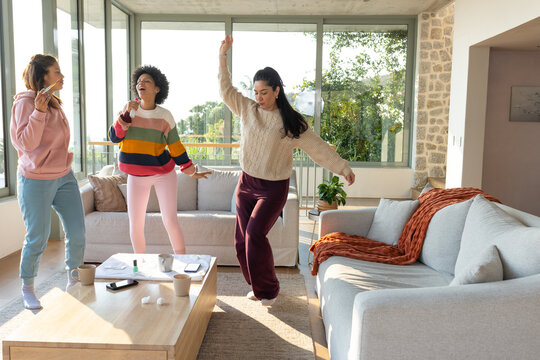 3 women friends wearing loungewear dancing and singing in living room near coffee table with phone