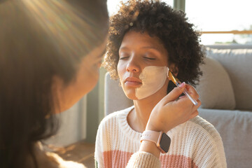 Diverse female friends sitting on sunlit couch, one applying cream mask with flat brush