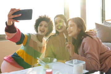 Diverse female friends sitting on sofa backlit by window, taking selfie with phone and mixing bowl