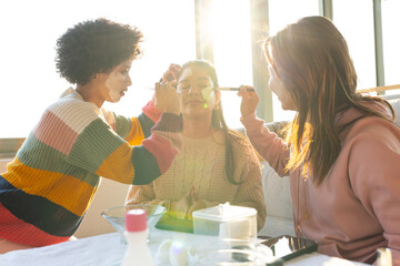Diverse female friends applying makeup on seated woman at lounge using brushes and green palette