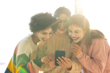 Diverse female friends leaning, wearing facial masks and sweaters, checking smartphone by window