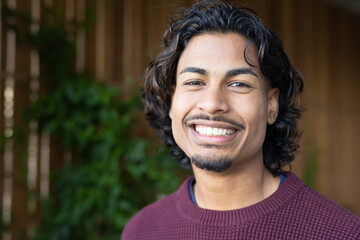 Man with wavy hair, goatee smiling outside by wood fence wearing burgundy sweater, copy space