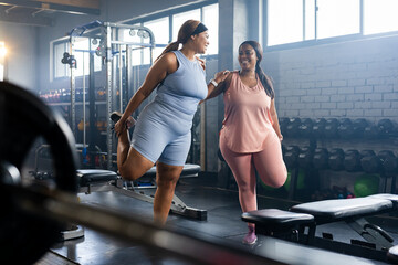Diverse mid-adult female friends stretching quads in gym wearing athletic clothes near dumbbells