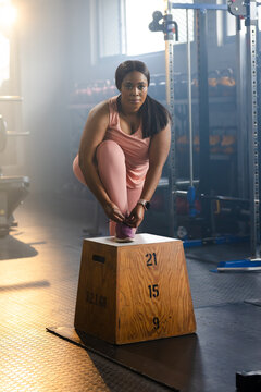 Mid-adult African American woman tying purple shoe on plyo box at gym kettlebells visible