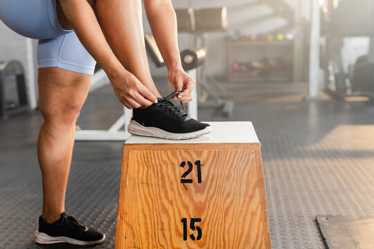 Hands and lower body are tying sneaker shoelace on plyo box in gym, stenciled 21 15