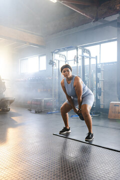 African American mid-adult woman lifting kettlebell in warehouse gym in sportswear by squat rig