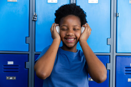 African teenage girl standing at blue school lockers in blue tee holding white headphones smiling