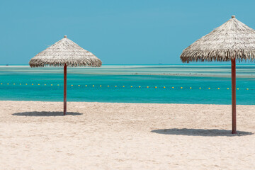 Sunny beach with two straw umbrellas over quiet turquoise water and soft sand, blue horizon