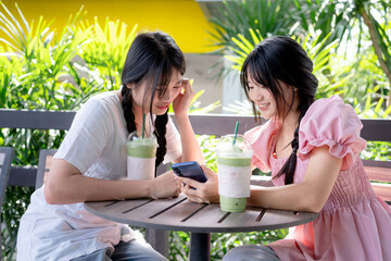 Two teenage girls sitting outdoors at a café with iced matcha drinks, using smartphones and enjoying a relaxed moment together in a green, natural setting.