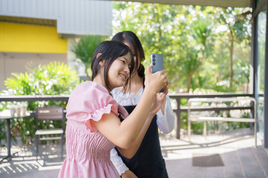 Two teenage girls taking a cheerful selfie together on an outdoor café terrace, enjoying friendship, sunlight, and a fun moment surrounded by greenery. - Powered by Adobe