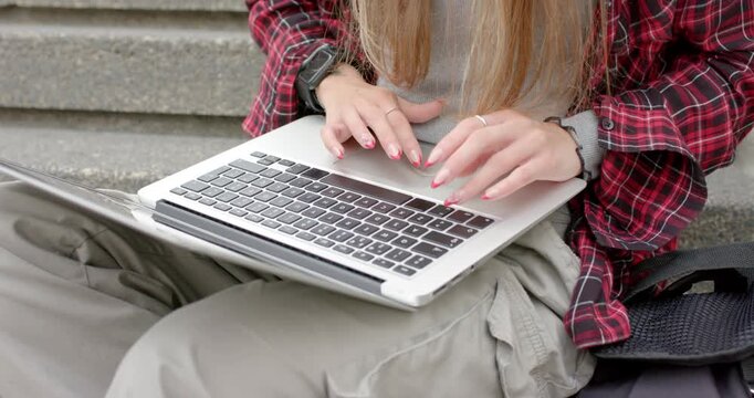Woman is settling onto concrete steps, placing silver laptop on lap and typing for remote work
