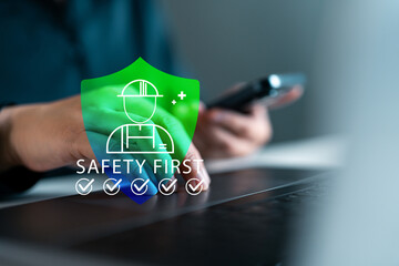 Workplace safety concept with digital technology. Hand using smartphone and laptop, green safety shield icon. Health and safety compliance in modern work environment.
