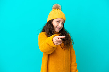 Little caucasian girl wearing winter hat isolated on blue background surprised and pointing front