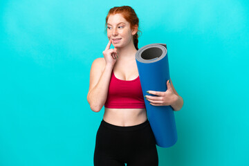 Young sport reddish woman going to yoga classes while holding a mat isolated on blue background...