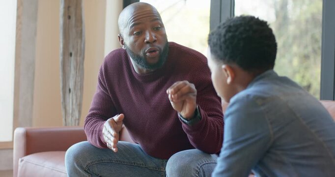Prompted by son leaning forward, African American father guiding with shrinking gestures on sofa