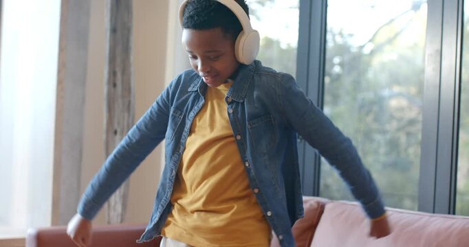 African American boy cueing music with white headphones, dancing in yellow T-shirt near glass door