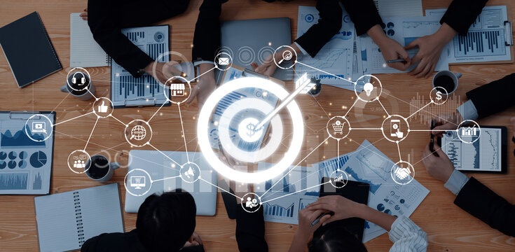 A top view of a business meeting featuring hands and documents with graphs. The central target graphic represents strategy and analytics in a corporate environment. Trope