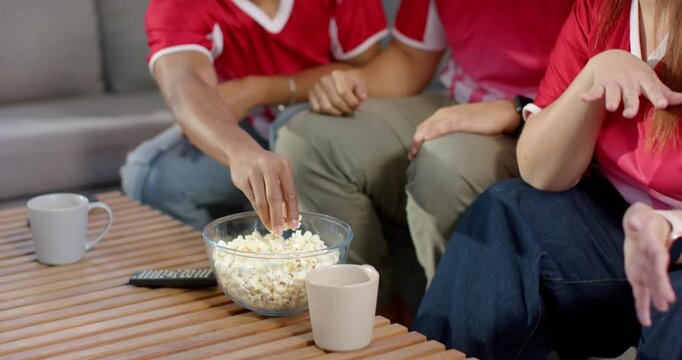Reacting to game, Diverse friends in red jerseys reaching into popcorn bowl, sharing in living room