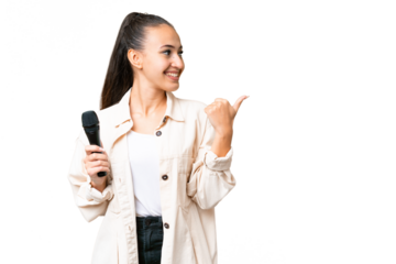 Young singer woman picking up a microphone over isolated chroma key background pointing to the side to present a product