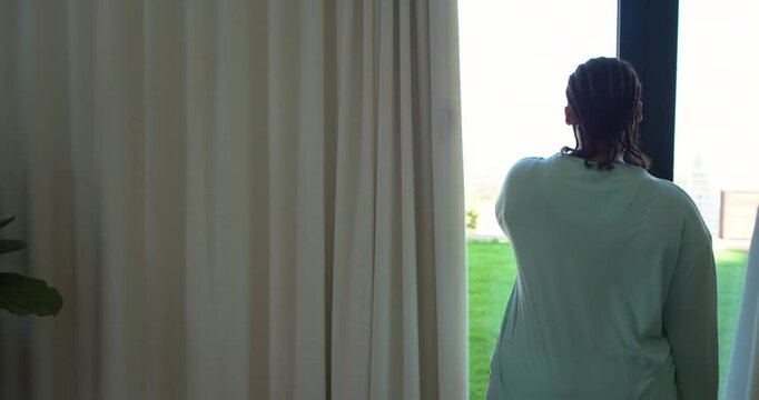 Woman is touching curtain rod and pulling beige curtain aside to view backyard lawn and sky