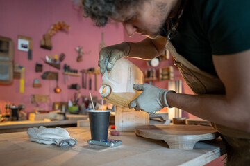 Craftsman carpenter in preparation process, working in own carpentry workshop, pouring required amount of epoxy resin for furniture detail and measures quantity on electronic scales on worktable.