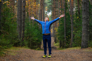 Young man standing on a forest trail with arms outstretched, breathing fresh autumn air and enjoying a peaceful, energizing moment of freedom and mindfulness while jogging