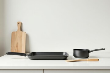 Modern Kitchen Utensils Sleek Black Frying Pan, Saucepan, and Wooden Spatula on a White Countertop Next to a Light Wood Cutting Board